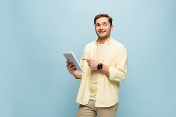 cheerful man in yellow shirt pointing with finger at digital tablet isolated on blue