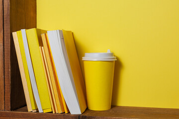 Books with yellow covers and a paper coffee cup sit on a brown old wooden shelf