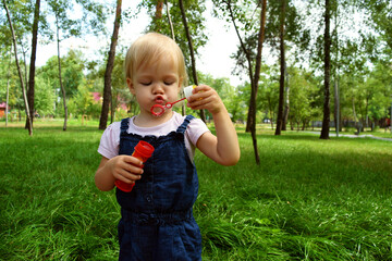 A little girl in a denim overalls blows bubbles in a green city park. Child's rest in nature