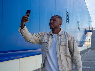 Fashionable black Spanish male using his smartphone against a blue wall