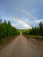 A scenic view of a double rainbow in the cloudy rainy sky over dense Oulanka National Park landscape with a rural path