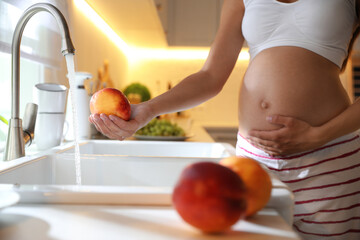 Young pregnant woman washing fresh sweet peach in kitchen, closeup. Taking care of baby health
