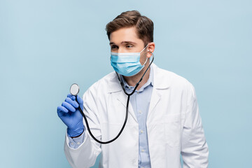 young doctor in medical mask and white coat holding stethoscope isolated on blue