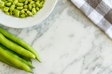 Overhead view of green organic bean pods and plate full of broad beans on marble table in kitchen. Rich and nutritious legumes. With copy space. 