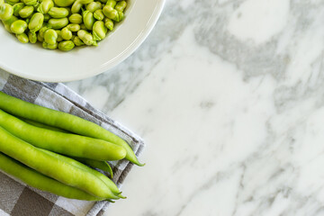 Overhead view of green organic bean pods and plate full of broad beans on marble table in kitchen. Rich and nutritious legumes. With copy space. 