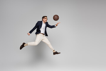 Fototapeta na wymiar full length of cheerful businessman playing basketball while levitating on grey