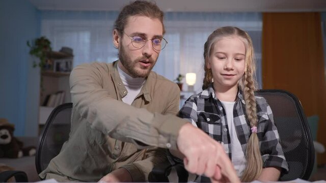 family looks in laptop and speaks at camera in internet, young dad in glasses and girl with cards in hands watching video lesson at table at home