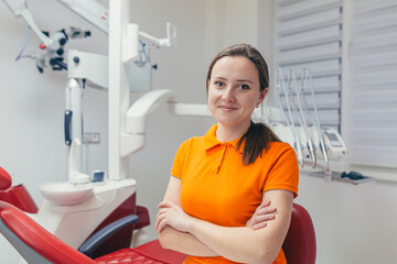 portrait young friendly female dentist smiling and looking at camera in a modern dental office. attractive woman specialist doctor sitting a chair with crossed arms on a white equipment background
