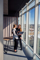 Fototapeta na wymiar Two successful female architects are talking about a joint project while standing at the window with a laptop. Young women economists dressed in formal attire talking during a break from work