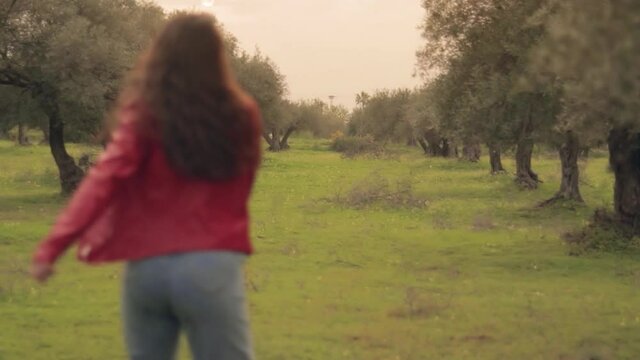 Mujer joven con chaqueta roja moviendo el cabello al viento en un prado verde con olivos y flores amarillas con el atardecer de fondo