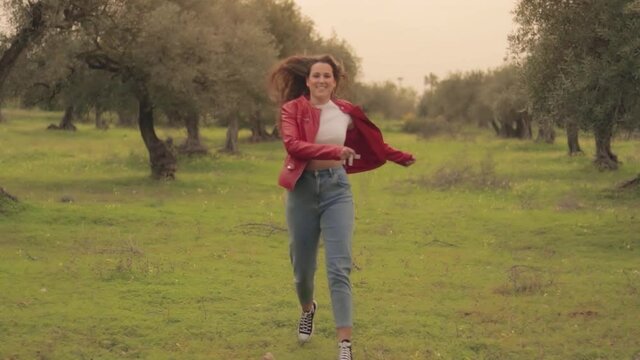 Mujer joven con chaqueta roja corriendo feliz en un prado verde con olivos y flores amarillas con el atardecer de fondo