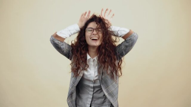 The crazy, amused young woman, with her hands above her head, emits horns and smiles broadly. Cretaceous in gray jacket and white shirt, with glasses posing isolated on a beige background in the