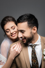 happy bride with closed eyes hugging happy muslim groom isolated on grey