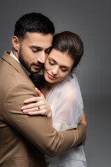 happy, interracial newlywed couple embracing with closed eyes isolated on grey