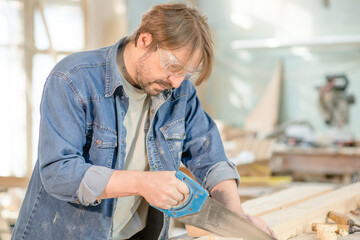 Carpenter sawing a board with a hand wood saw in carpentry workshop