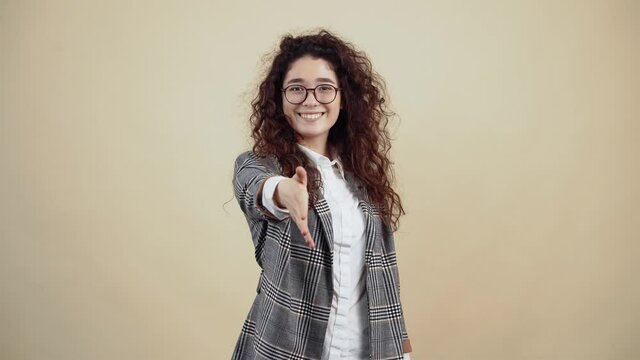 The positive young woman with a wide smile extends her hand in front of her to get to know her. Cretaceous in gray jacket and white shirt, with glasses posing isolated on a beige background in the