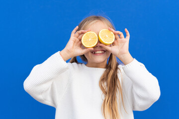 Overjoyed blond haired girl in white sweater smiling while covering eyes with bright lemon slices isolated on blue background. High quality photo
