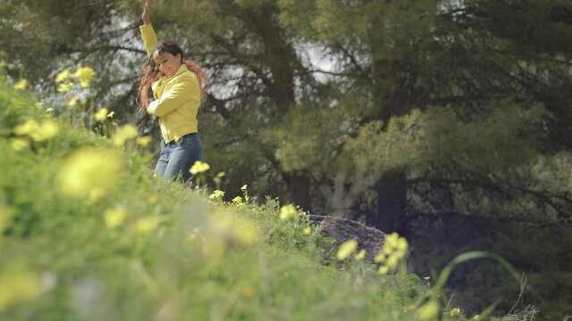 mujer joven con chaqueta amarilla bailando de felicidad en un bosque con un prado con flores amarillas en España