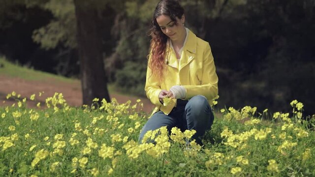 Mujer joven sonriendo y jugando con una flor en un prado con flores amarillas en un bosque con &aacute;rboles