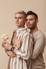 Minimal waist up portrait of young gay couple embracing and holding flowers while posing against neutral beige background in studio