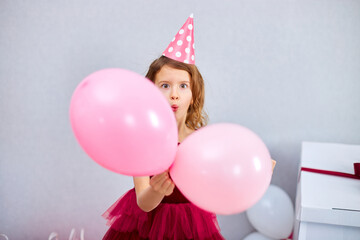 Fototapeta na wymiar Cute, Joyful little girl in pink dress and hat play with balloons at home birthday party