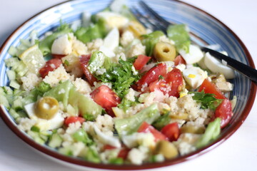 Salad with cuscus, lattice, tomato, cucumber, lemon, olives, onion, parsley, egg, olive oil in blue plate with fork on white background