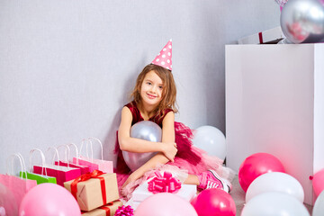 Little girl in pink dress and birthday hat sitting at the floor with many present gift box, balloons
