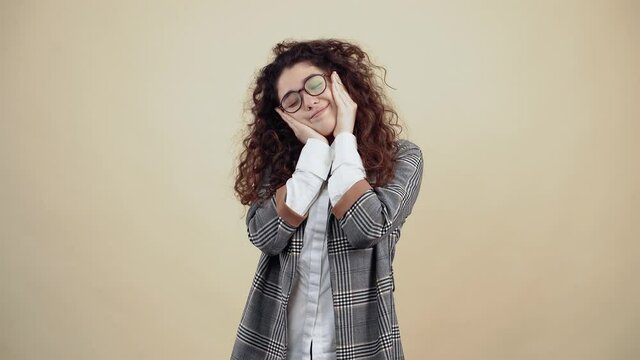 The young woman with her eyes closed, puts her hand on her face and with her big cheeks fools herself. Cretaceous in gray jacket and white shirt, with glasses posing isolated on a beige background in