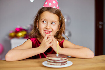 A little girl is wearing a birthday hat makes a wish , looking at a Birthday Cake