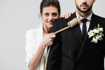 Smiling bride holding slip knot on neck of groom in suit isolated on grey