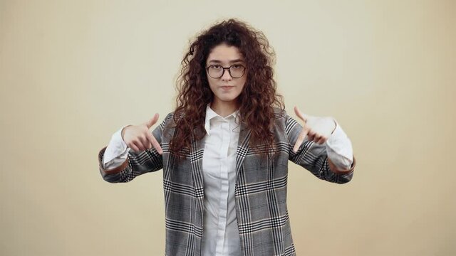 Hey subscribe here, the resentful young woman looks with both fingers down like a gesture. Cretaceous in gray jacket and white shirt, with glasses posing isolated on a beige background in the studio