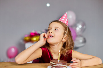 A little girl is wearing a birthday hat makes a wish , looking at a Birthday Cake