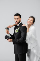 Smiling bride taking money from arabian boyfriend with wallet isolated on grey