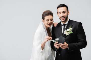 Smiling bride looking at wallet with money in hands of muslim groom isolated on grey