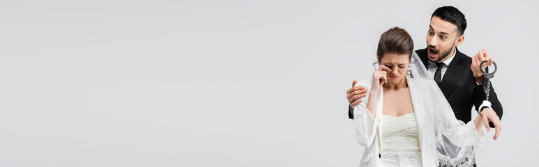 Excited arabian groom hugging crying bride in handcuffs isolated on grey, banner