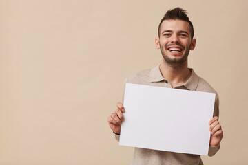Minimal waist up portrait of carefree young man holding blank white sign and smiling at camera while posing against beige background in studio, copy space
