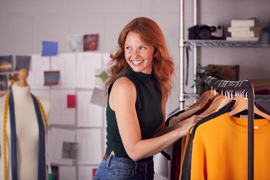 Female Owner Of Fashion Business Checking Clothing Samples On Rail In Studio