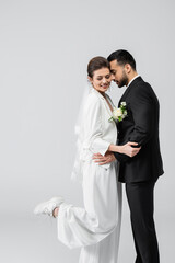 Muslim groom in suit hugging cheerful bride isolated on grey