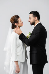 Side view of muslim groom touching neck of smiling bride isolated on grey