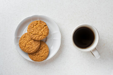 White cup with coffee and white saucer with oatmeal cookies on a white table, top view. Morning routine concept.
