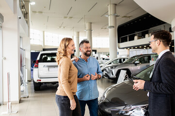 Young car salesman showing to mid-age couple new automobile at dealership salon.