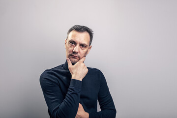 Fototapeta premium A white middle aged man of European appearance with gray in his hair and a slightly unshaven face in a black turtleneck looks proudly at the camera supporting his head by the chin