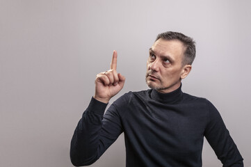 A white man of European appearance middle aged with gray in his hair and a slightly unshaven face in a black turtleneck thoughtfully points his index finger up