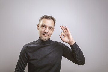 A white man of European appearance middle aged with gray in his hair and a slightly unshaven face in a black turtleneck cheerfully shows OK with his left hand
