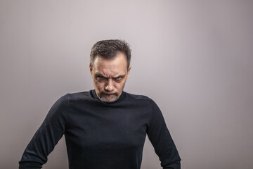 A middle aged white man of European appearance with gray in his hair and a slightly unshaven face in a black turtleneck frowned menacingly