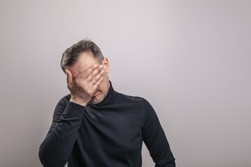 A white middle aged man of European appearance with gray in his hair and a slightly unshaven face in a black turtleneck covered his face with his hand in despair