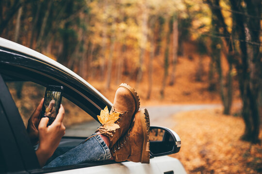 Woman takes picture of bright yellow leaf with mobile phone sticking legs out of car window in picturesque autumn forest