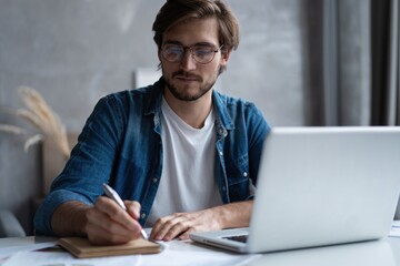 Serious young businessman with the glasses sitting and using laptop at home office and taking notes.