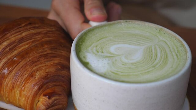 Mug of matcha tea latte served with fresh croissants. Hand taking a mug away
