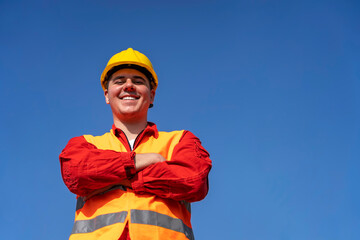Young Worker in Red Coveralls, Reflective Clothing and Yellow Hardhat Standing Against Blue Sky Background